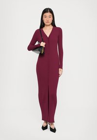 Ribbed, burgundy long-sleeve dress with a V-neck, button-down front, and front slit. Worn with a black handbag and pointed shoes.