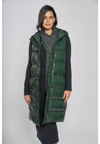 Bezrekawnik zielonej puchowej kamizelki z połyskującym wykończeniem, pikowanym wzorem, z kapturem, zestawiony ze szarym szalikiem i długą czarną sukienką.
