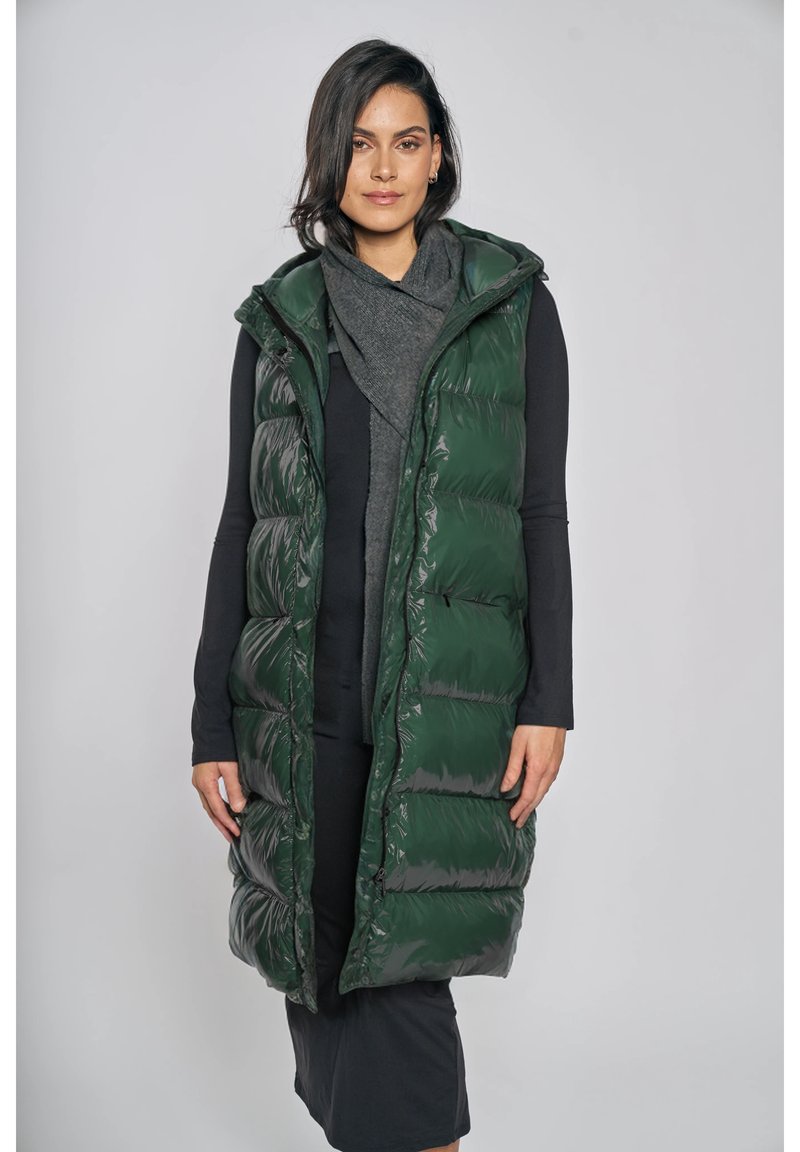 Bezrekawnik zielonej puchowej kamizelki z połyskującym wykończeniem, pikowanym wzorem, z kapturem, zestawiony ze szarym szalikiem i długą czarną sukienką.