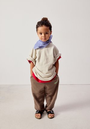 Otrok z lasmi v figi, oblečen v bež srajco z zavihanimi rokavi, rdečo spodnjo majico, modro karo rutko okoli vratu, rjave hlače in črne sandale stoji.
