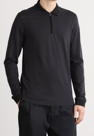 Homme portant un polo noir à manches longues avec une courte fermeture éclair sur le devant et un pantalon noir, mains partiellement dans les poches.