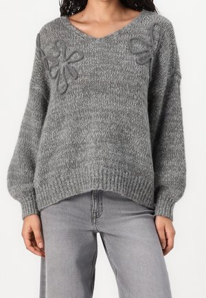Femme portant un pull en maille gris ample avec des détails floraux en fil bouclé près des épaules et un jean gris clair à jambes droites.