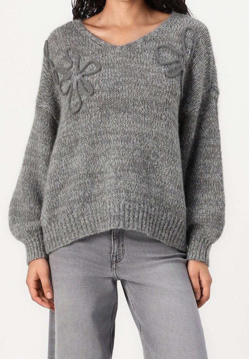 Femme portant un pull en maille gris ample avec des détails floraux en fil bouclé près des épaules et un jean gris clair à jambes droites.