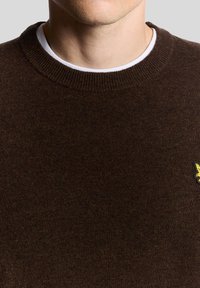 Καφέ πουλόβερ από πλεκτό ύφασμα με ριμπ λαιμόκοψη. Διαθέτει μικρή κεντημένη λεπτομέρεια στην κάτω γωνία.