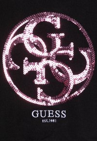 Czarna koszulka z dużym, błyszczącym różowym logo z cekinów oraz białym haftowanym napisem "GUESS EST. 1981" poniżej.