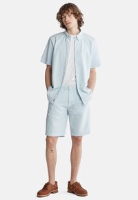 Lys blå kortærmet skjorte med knapper, hvid t-shirt under, parret med lys blå mønstrede shorts og brune snørrede sko.