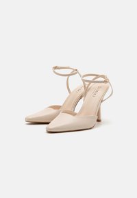 Chaussures à talons hauts beiges avec un bout pointu et des brides de cheville. Matériau en simili cuir lisse avec un détail de couture subtil. Talon de hauteur moyenne.