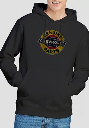 Eine lächelnde Person trägt einen schwarzen Hoodie mit einem abgenutzten gelben und roten Chevrolet-Logo, auf dem "Echte Teile" steht, kombiniert mit blauen Jeans.