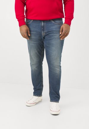 Personne portant un jean en denim bleu, un t-shirt à manches longues rouge et des baskets blanches, debout devant un fond uni.