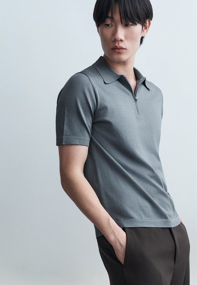 Jeune homme aux cheveux noirs portant un polo gris à manches courtes zippé et un pantalon foncé, regardant vers sa gauche avec les mains dans les poches.