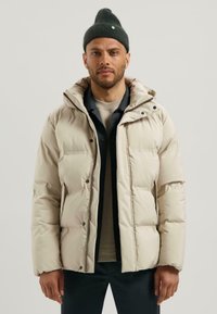 Beige gewatteerde jas met een ritssluiting aan de voorkant, capuchon en drukknoopsluiting. Zachte textuur, gequilt ontwerp, gedragen over een donkere shirt en lichte trui.