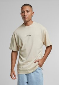 Beige bomulds t-shirt med en afslappet pasform og korte ærmer. Har et lille sort logo "AnotherCotton" centreret på brystet.