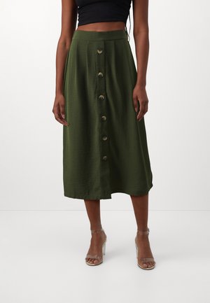 Vrouw draagt een olijfgroene midi-rok met hoge taille en knopen aan de voorkant, een zwart cropped topje en transparante sandaalhakken, tegen een effen achtergrond.