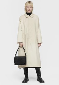 Cappotto oversize beige con colletto, dolcevita nero, stivali in pelle nera e borsa in pelle nera con il logo "TOUS" in rilievo.