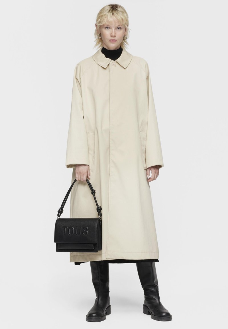 Cappotto oversize beige con colletto, dolcevita nero, stivali in pelle nera e borsa in pelle nera con il logo "TOUS" in rilievo.