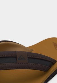 Κοντινή λήψη καφέ και μαύρης σαγιονάρας με υφασμένους ιμάντες και λογότυπο στο ταμπά πέλμα πάνω σε λευκό φόντο.