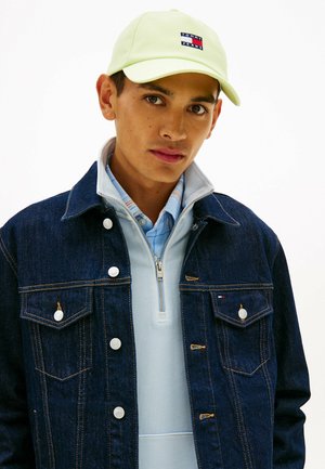 Jonge man met een limegroene Tommy Jeans pet, een lichtblauwe zip-up trui in laagjes, een geruit overhemd en een donker denim jack met drukknoopsluiting.