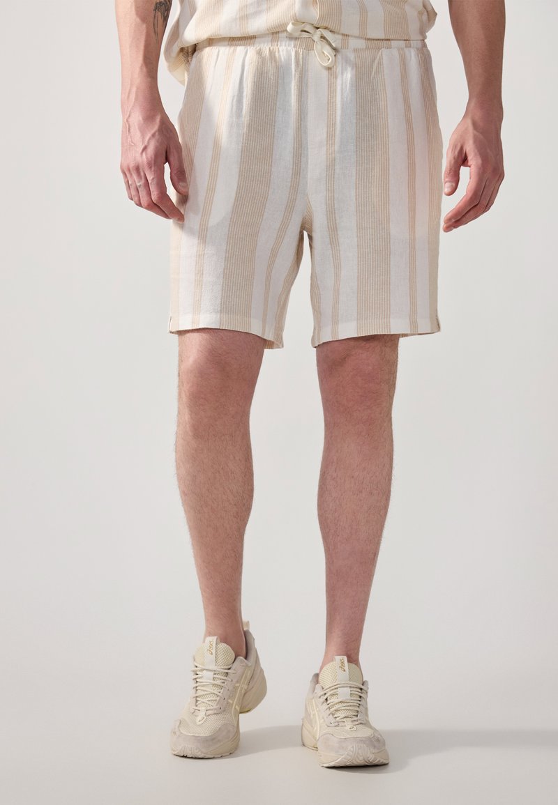 Pantaloni scurți bej și albi cu dungi, cu talie elastică și șnur de ajustare. Stil casual, cu o croială lejeră, asortați cu adidași ușori.