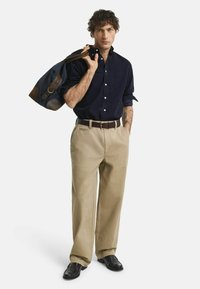 Marineblauwe overhemd, beige wijde broek, bruine leren riem en donkere instappers. Draagt een blauwe tas met bruine leren accenten.