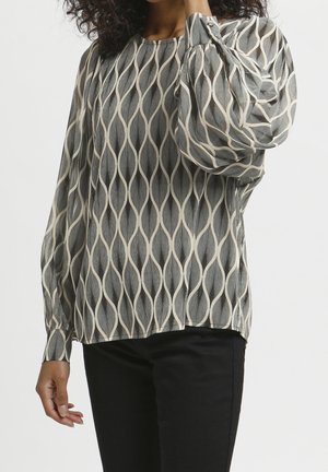 Kvinde iført en langærmet bluse med et sort og beige bølgemønster og sorte bukser, stående med den ene hånd, der berører hendes nakke.