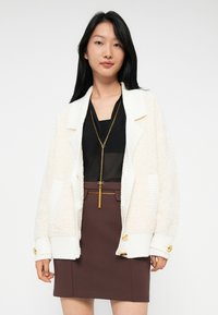 Woman wearing a white textured jacket, black sheer top, brown skirt with gold-chain detail, and long gold necklace, standing against a white background.