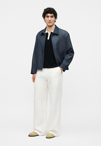 Junger Mann trägt eine marineblaue Jacke, schwarz-weißes Poloshirt, weite weiße Hose und beige Loafers, steht vor einem schlichten weißen Hintergrund.