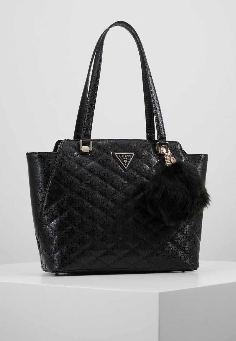 Sac fourre-tout noir matelassé avec motif logo, texture lisse, garnitures dorées, double poignée, et un pompon en fourrure noire amovible.