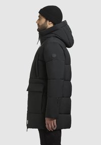 Black puffer jacket with a hood, featuring a zippered pocket, a large front pocket, and soft, quilted texture. Styled with a matching beanie.
