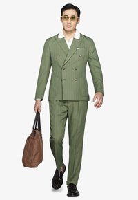 Mand iført en grøn nålestribet dobbeltbrystet jakkesæt, hvid skjorte, solbriller med gult glas, sorte sko og bærende en brun indkøbstaske.