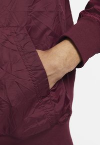 Burgundijasta jakna z nagubano teksturo, elastičnimi manšetami in stranskim žepom, izdelana iz lahkega sintetičnega materiala.