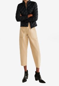 Veste matelassée noire avec fermeture éclair, pantalon croisé beige ajusté avec poches, et bottines noires à bout pointu et talon épais.