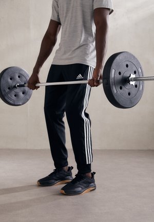 Schwarze Gewichtheberstange mit runden Gewichten, getragen von einer Person in einem grauen T-Shirt und schwarzen Trainingshosen mit weißen Streifen, schwarzen Sportschuhen.