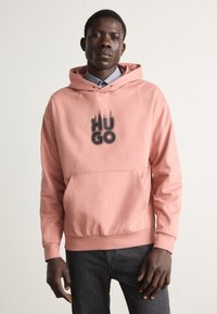 Sudadera rosa con un bolsillo frontal, que presenta "HUGO" en texto de degradado negro. Material suave con capucha con cordones y puños acanalados. Combinada con jeans oscuros.