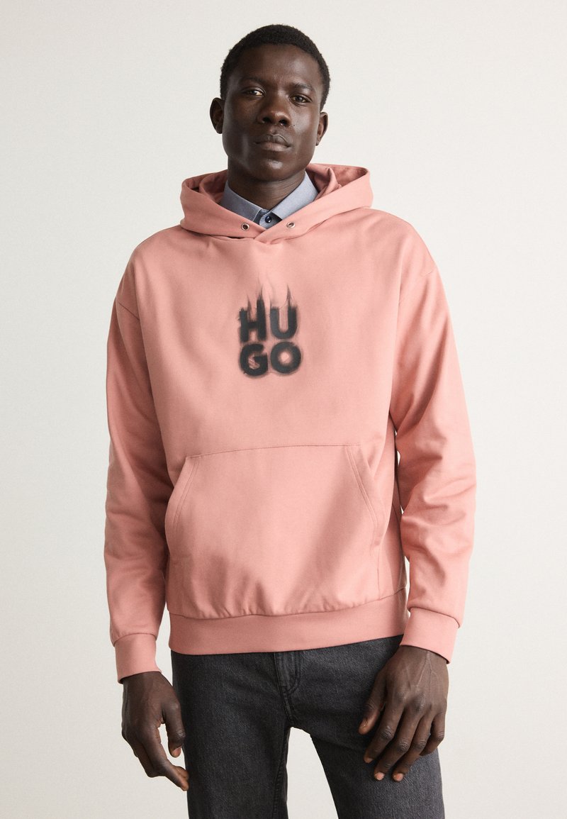 Sudadera rosa con un bolsillo frontal, que presenta "HUGO" en texto de degradado negro. Material suave con capucha con cordones y puños acanalados. Combinada con jeans oscuros.