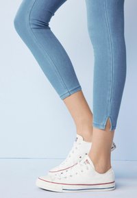 Jean skinny bleu clair avec ourlet retroussé et fentes latérales, accompagné de baskets en toile blanches avec des accents rouges et bleus.