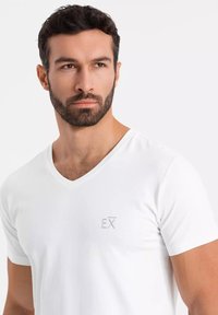 Biała bawełniana koszulka typu v-neck z subtelnym szarym logo po lewej stronie klatki piersiowej. Krótkie rękawy i miękka tekstura. Prosty i minimalistyczny design.