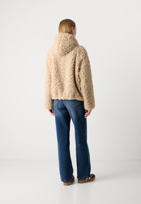 Person som har på seg en beige fluffy hettejakke, blå jeans og brune sko, stående med ryggen til mot en ensfarget hvit bakgrunn.