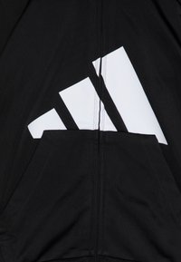 Sweat-shirt noir avec un grand logo blanc à trois bandes. Il présente une poche avant, un tissu texturé et des coutures visibles le long de la fermeture éclair.