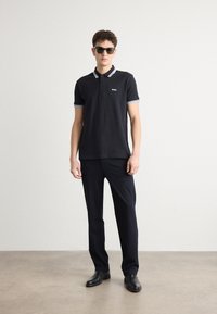 Crna polo majica s ovratnikom u bijelim i plavim prugama, izvezeni logo na prsima, uparen s tamnim hlačama i crnim cipelama. Model nosi sunčane naočale.