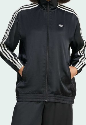 Osoba nosząca czarną kurtkę sportową Adidas z białymi paskami na rękawach i rękami w kieszeniach, widoczna od szyi do bioder.