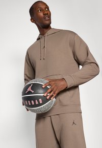 Hnedá mikina s kapucňou a sťahovacou šnúrkou, s malým logom na hrudi. Osoba drží čierno-sivý basketbal s logom a červenými akcentmi.