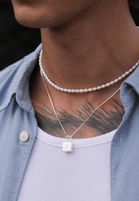 Sølv lås vedhæng på en tynd kæde over en hvid ribbet skjorte, sammen med en hvid perle choker, mod en baggrund af mørk tekstur.