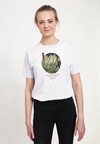 Bílé bavlněné tričko s kruhovým motivem bambusu v zelených tónech a červených akcenty v uměleckém stylu. Černé džíny na sobě.