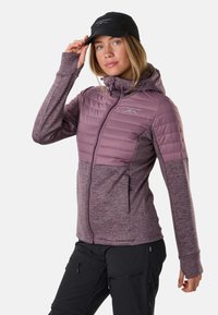 Frau trägt eine violette gesteppte und gestrickte Zip-Up-Jacke mit Kapuze, schwarze Mütze und schwarze Cargohosen, richtet die Mütze mit der rechten Hand.