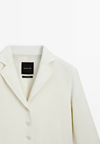 Helle cremefarbene maßgeschneiderte Blazerjacke mit Reverskragen, fünf Knöpfen und weicher Textur. Das Innenetikett zeigt den Markennamen und Produktionsdetails.