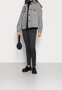 Veste texturée grise avec poches avant, pull noir, pantalon skinny gris foncé, et mocassins chunky noirs. Tenant un sac à main noir tressé.