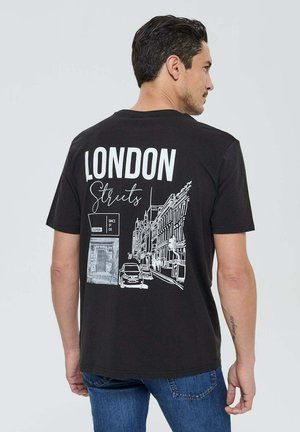Homme portant un t-shirt noir avec une illustration de rue de Londres en blanc et du texte, associé à un jean bleu, vu de dos.