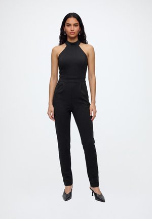 Femme debout sur fond blanc, portant une combinaison noire sans manches et des chaussures à talons noirs, faisant face à l'avant avec les bras le long du corps.