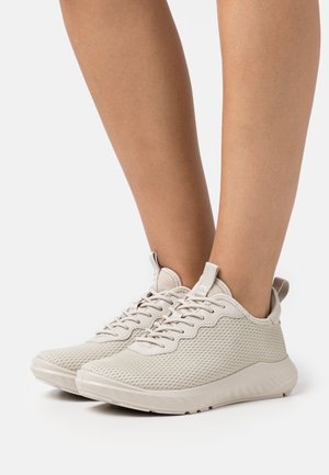 Baskets en mesh beige clair avec lacets portées aux pieds sur un fond blanc Uni.