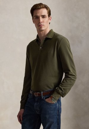 Mann trägt ein olivgrünes langärmeliges Poloshirt, blaue Jeans, braunen Gürtel und eine Armbanduhr, steht vor einem schlichten neutralen Hintergrund.